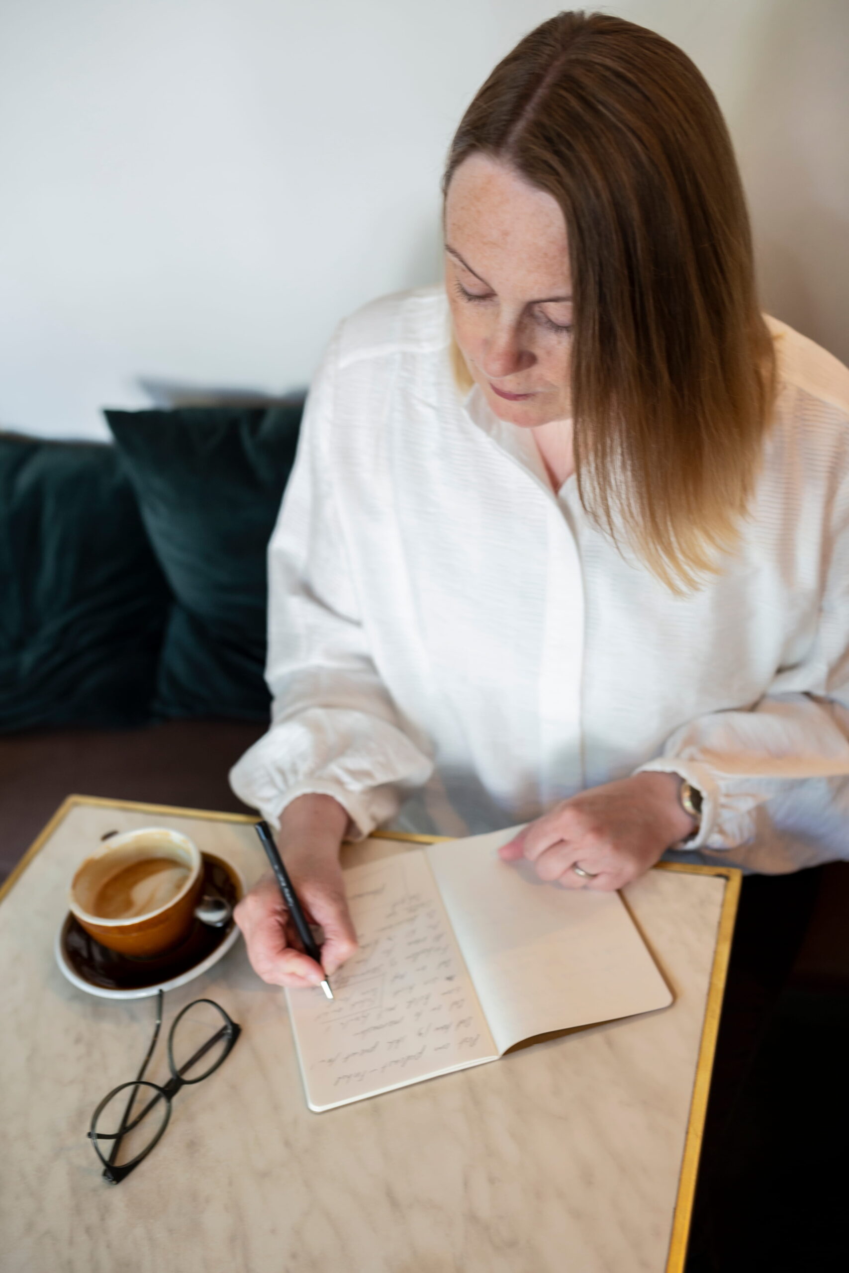 Lene stampe sidder ved et cafébord med en kopkaffe og skriver i en notesbog